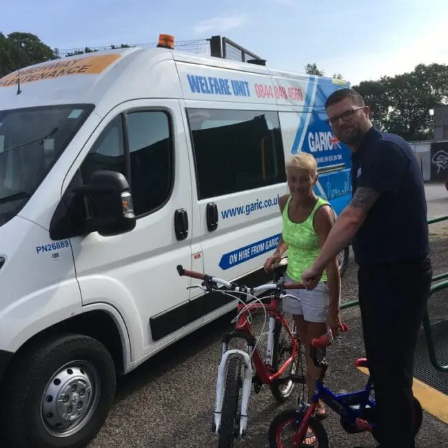 A man and woman stand with bicycles next to a white welfare unit van on a sunny day.