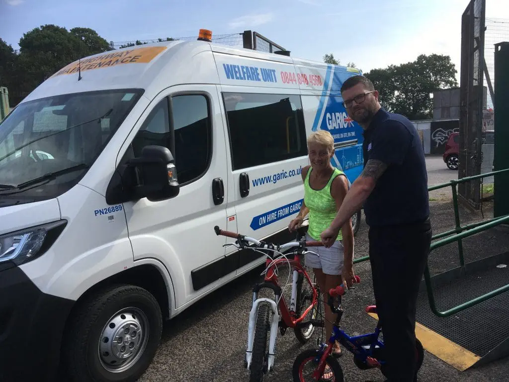 A man and woman stand with bicycles next to a white welfare unit van on a sunny day.