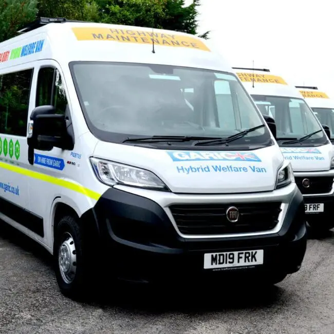 A row of highway maintenance hybrid welfare vans parked beside a road.