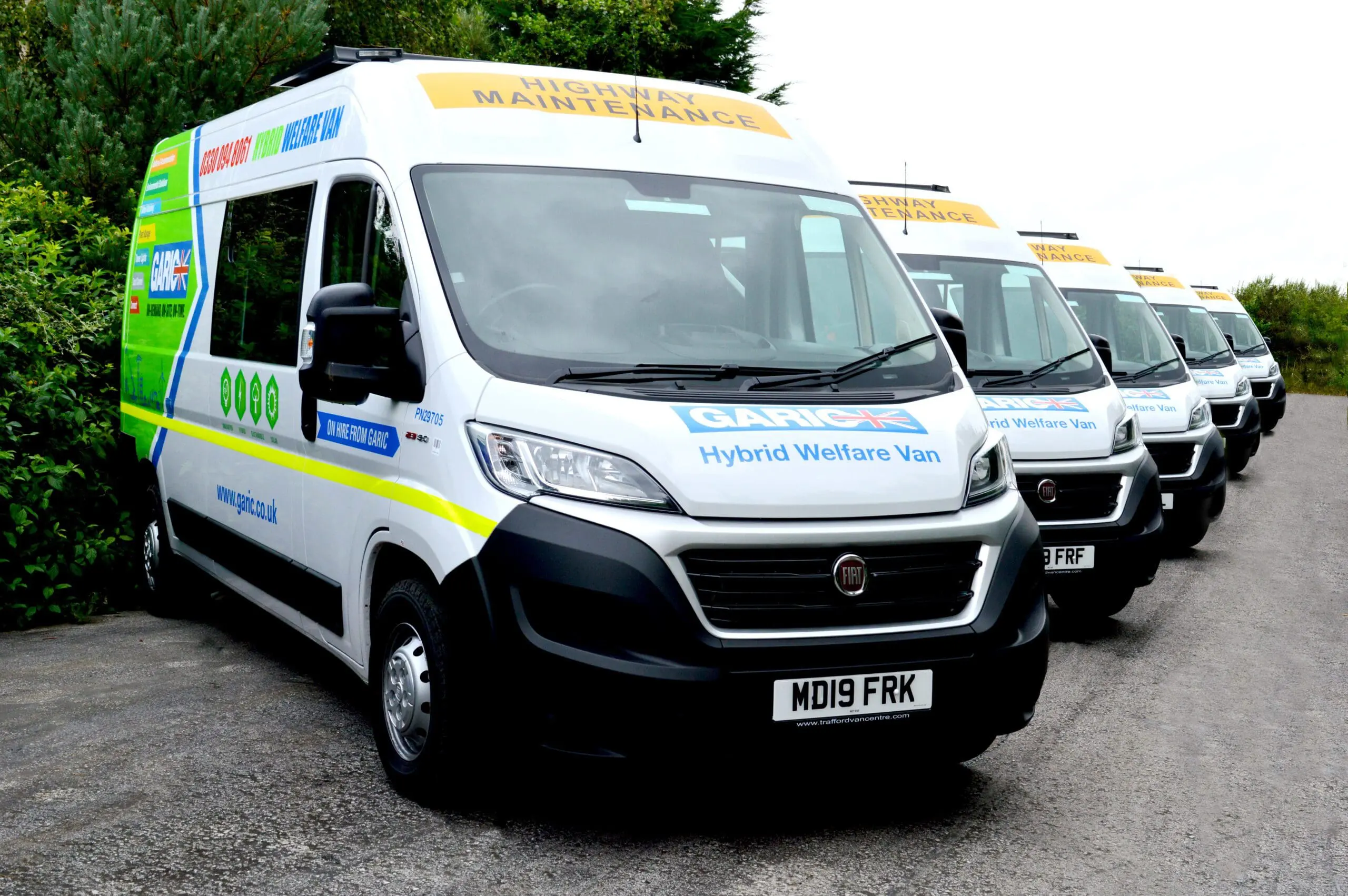 A row of highway maintenance hybrid welfare vans parked beside a road.