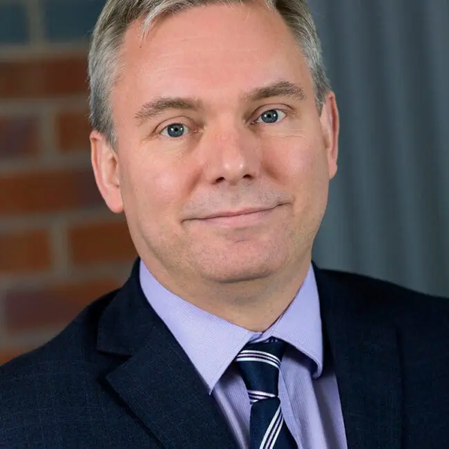 A man in a suit and striped tie poses in front of a brick and gray background, smiling slightly.