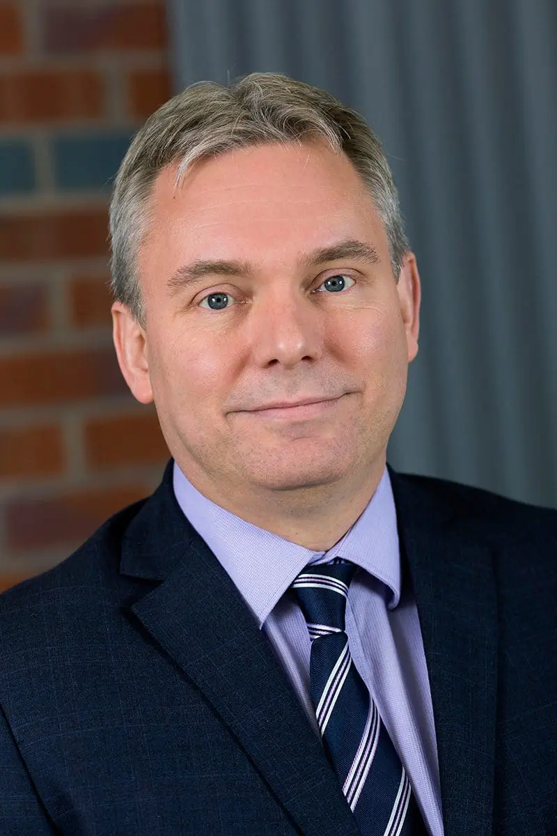 A man in a suit and striped tie poses in front of a brick and gray background, smiling slightly.