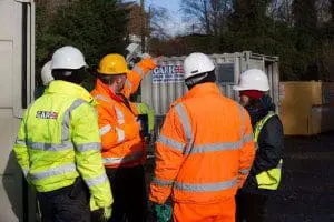 Four construction workers in high-visibility jackets and helmets talk outside near a metal container.
