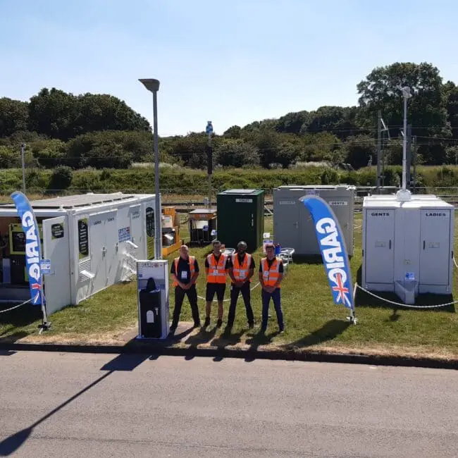 Four people in high-visibility vests stand in front of portable toilets and cabins on a sunny day.