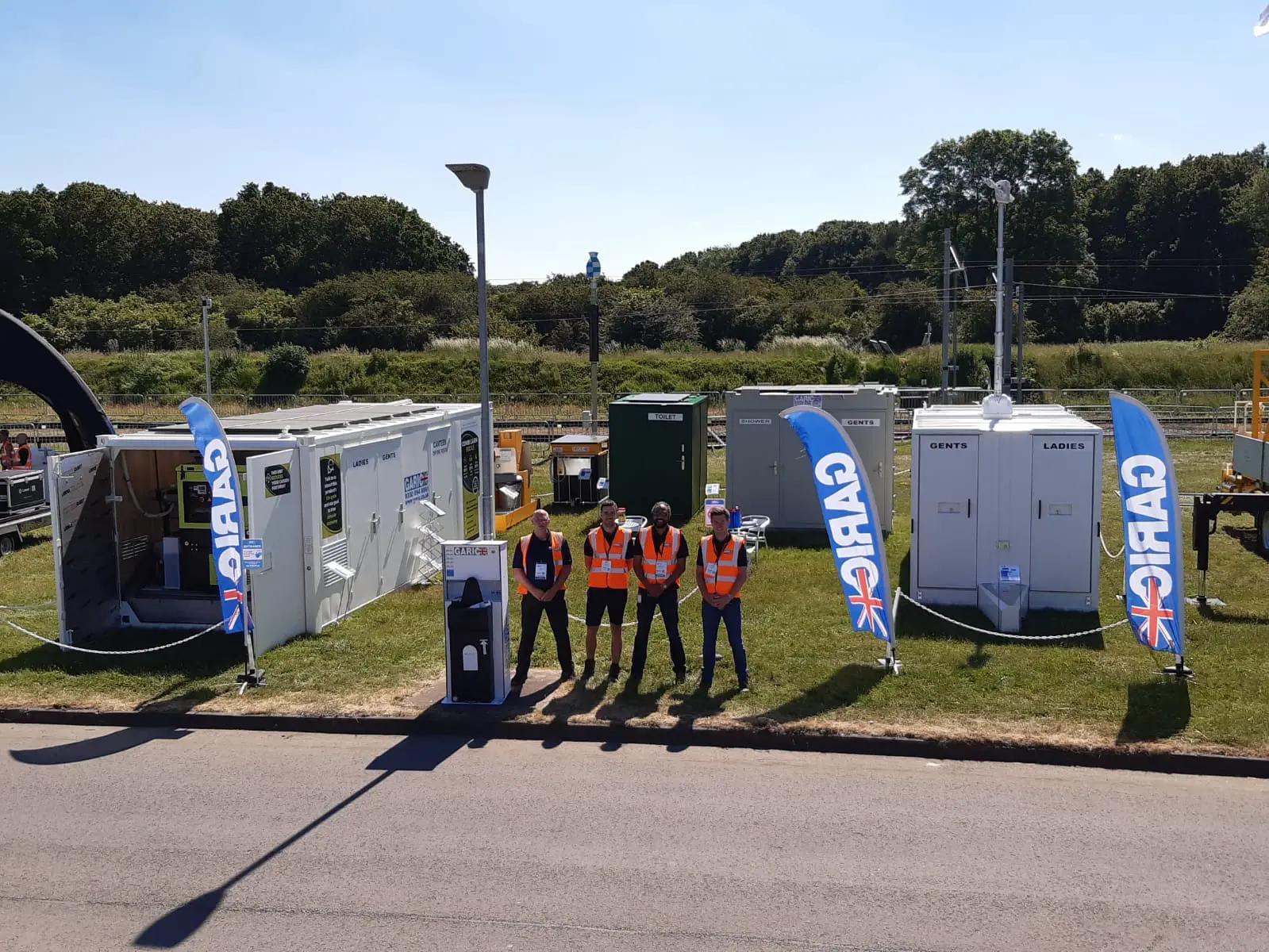 Four people in high-visibility vests stand in front of portable toilets and cabins on a sunny day.