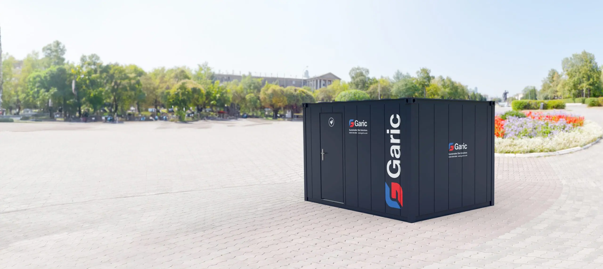 A 2+1 Toilet Block 12 in dark gray is situated on a paved area within a park, surrounded by trees and flower beds.