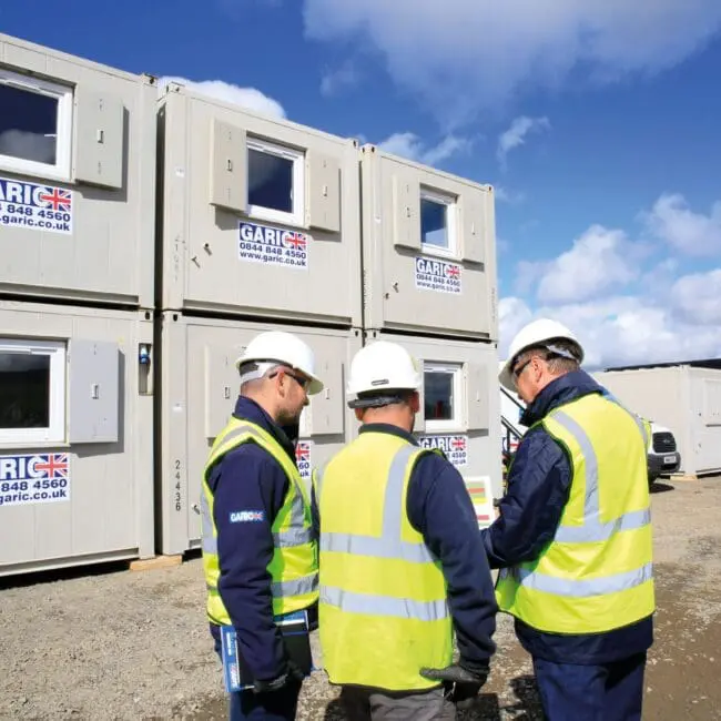 Three construction workers wearing high-visibility vests and helmets stand near stacked portable cabins.