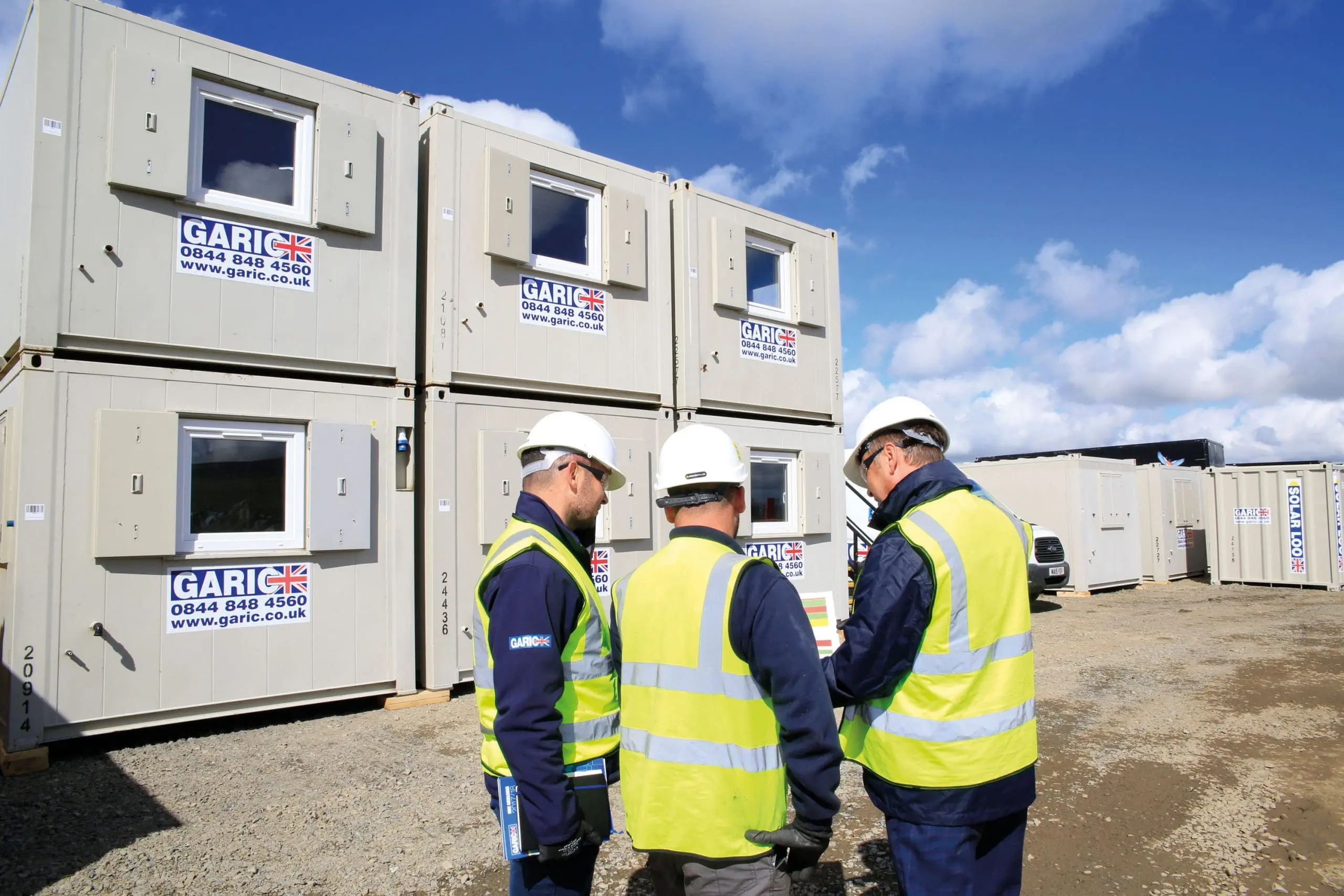 Three construction workers wearing high-visibility vests and helmets stand near stacked portable cabins.