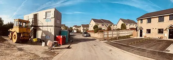 A suburban street with new houses, construction equipment, and temporary site offices under a blue sky.