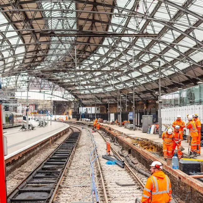 Railway workers in orange vests repair tracks under a large glass roof at a train station.