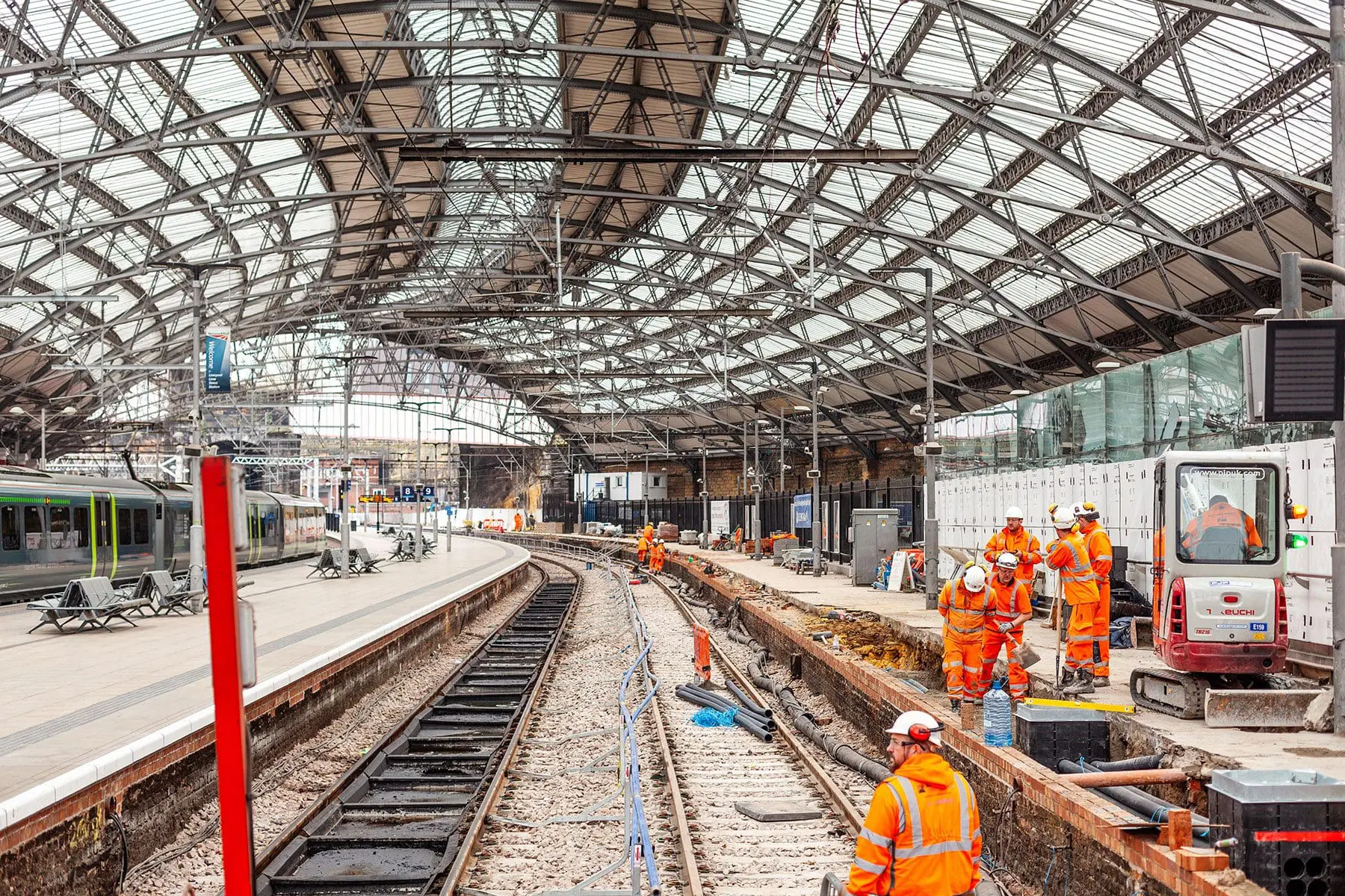 Railway workers in orange vests repair tracks under a large glass roof at a train station.