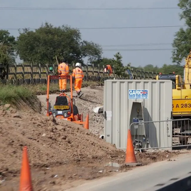 Construction workers in orange vests working near machinery and cones on a roadside site.