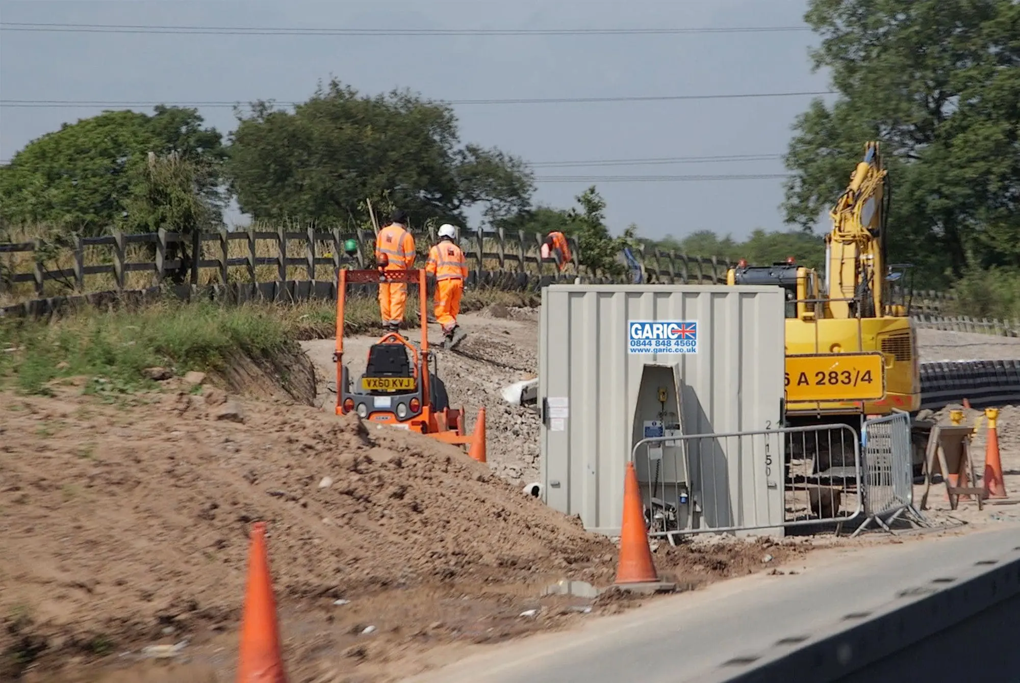 Construction workers in orange vests working near machinery and cones on a roadside site.