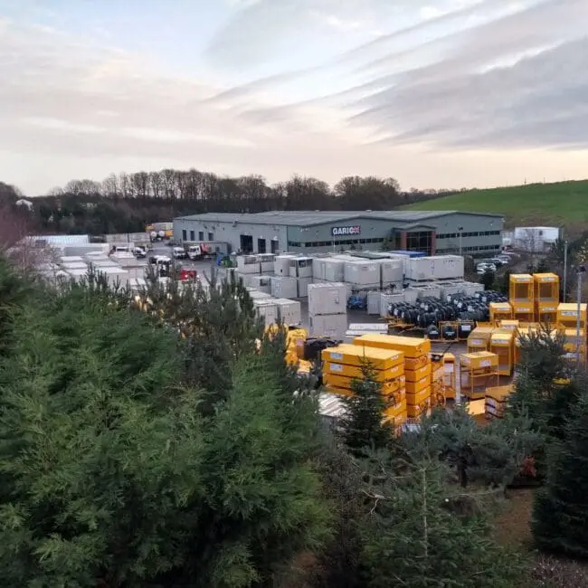 Industrial facility with stacks of yellow and white containers, surrounded by trees and fields under a cloudy sky.