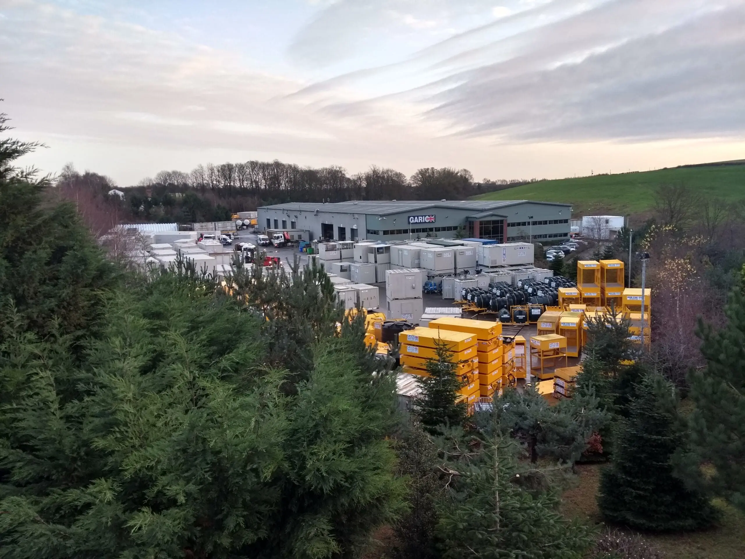 Industrial facility with stacks of yellow and white containers, surrounded by trees and fields under a cloudy sky.