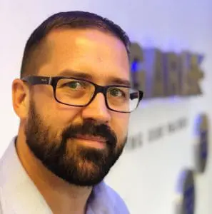 Man with dark hair, beard, and glasses smiling, standing indoors near a blurred wall sign.