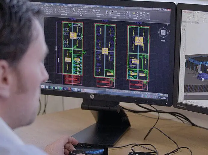 A person works on architectural floor plans displayed on dual computer monitors using design software.
