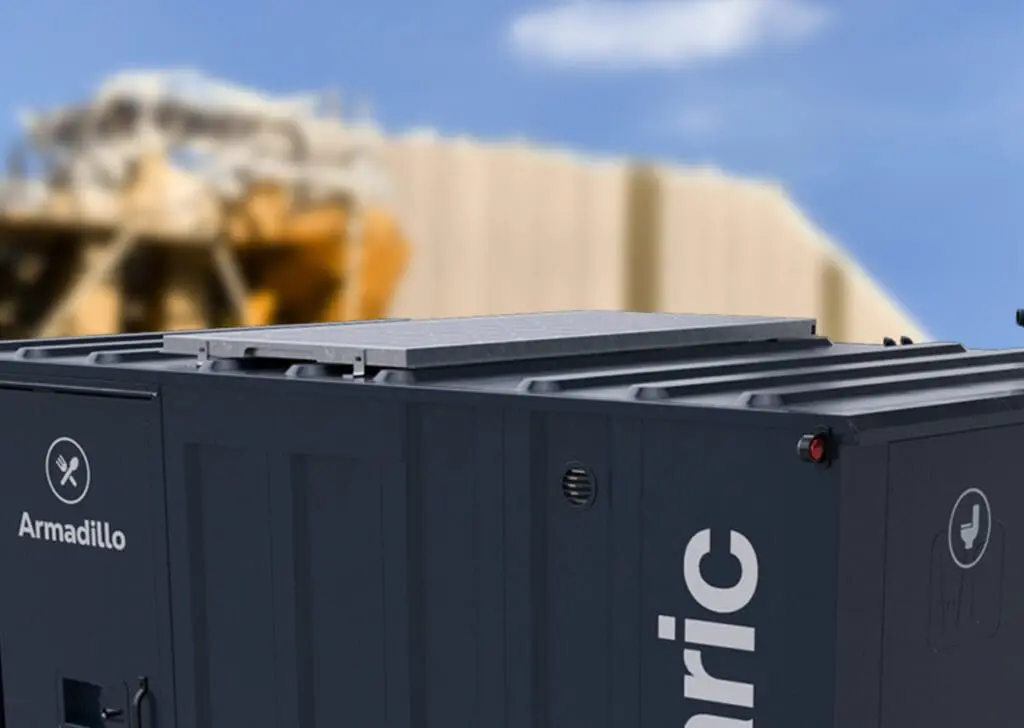 A dark gray Armadillo 12 Eco container stands with a blurred industrial structure in the background.