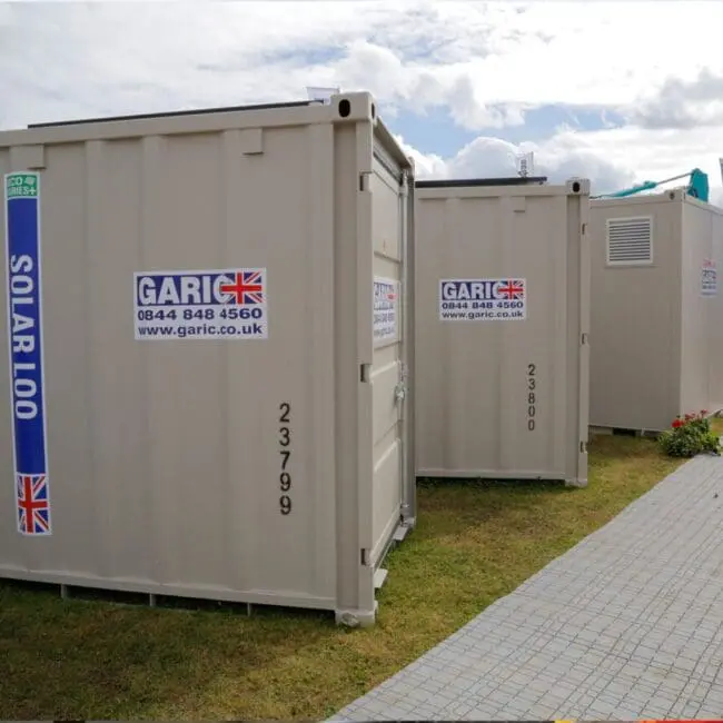 Portable toilets labeled Solar Loo and Garic are placed outdoors on a grassy area near a walkway.