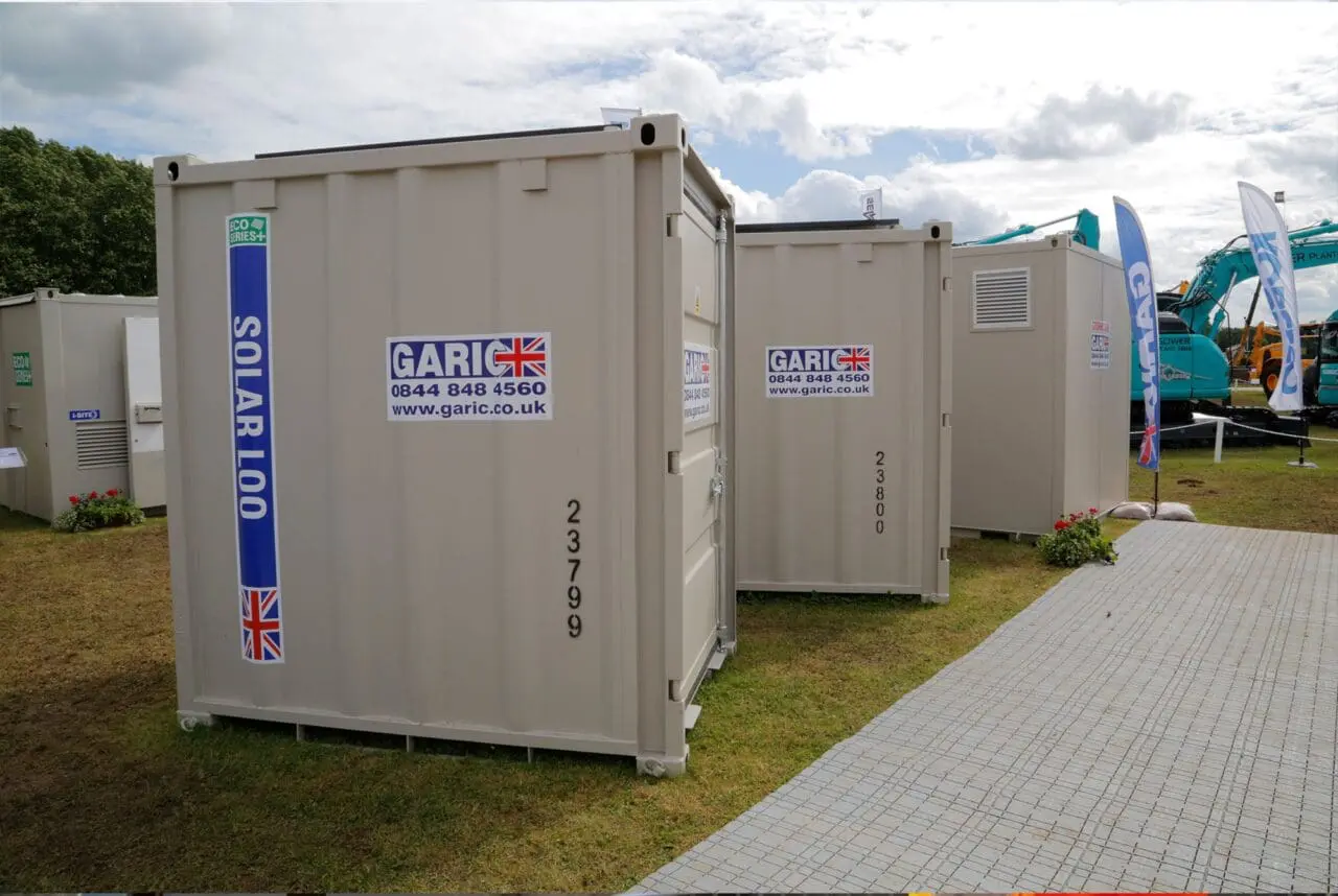 Portable toilets labeled Solar Loo and Garic are placed outdoors on a grassy area near a walkway.