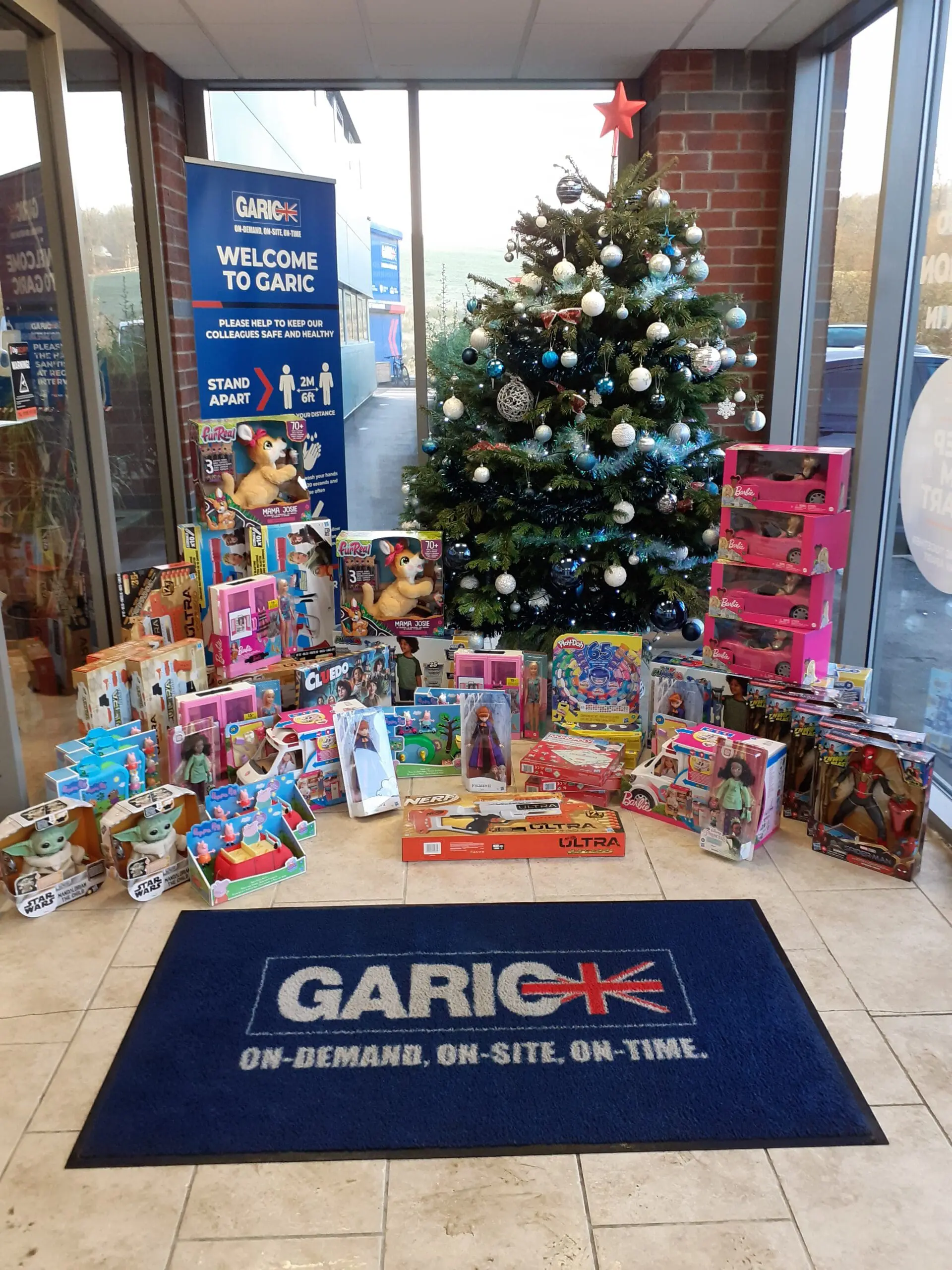 Christmas tree with ornaments surrounded by various donated toys inside an office lobby with Garic branding.