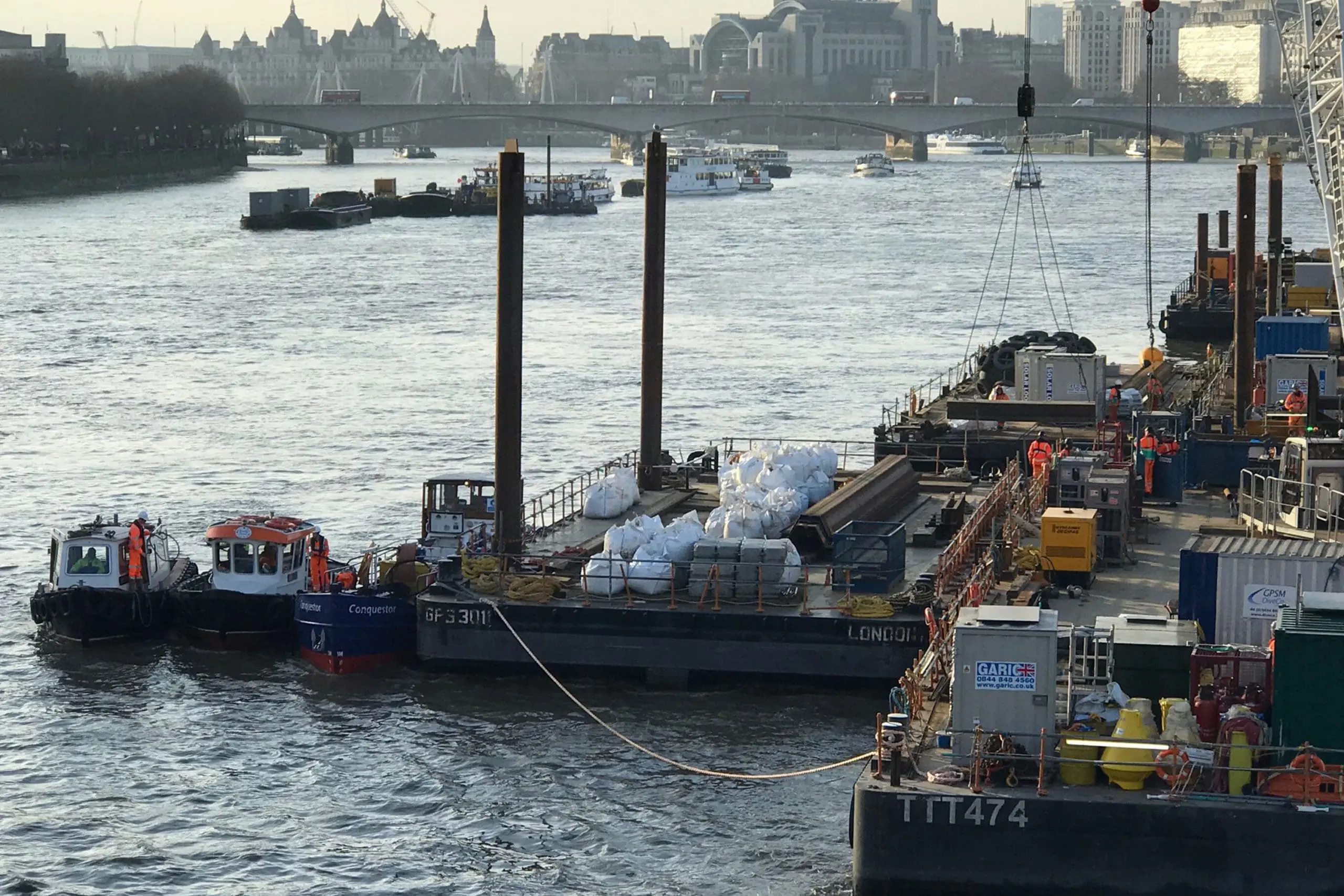 Barges and workers on the River Thames with construction materials and equipment, cityscape in background.