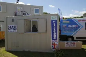 A portable office cabin and van with GARIC branding, set outdoors on grass under a blue sky.