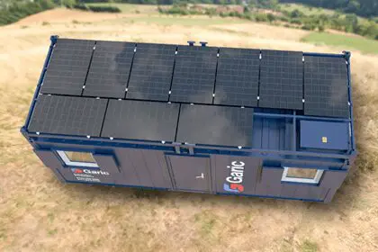 A blue container building with solar panels on its roof, situated on a grassy field.