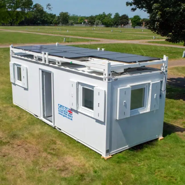 White portable container unit with rooftop solar panels, placed on grass in an open, sunny park area.