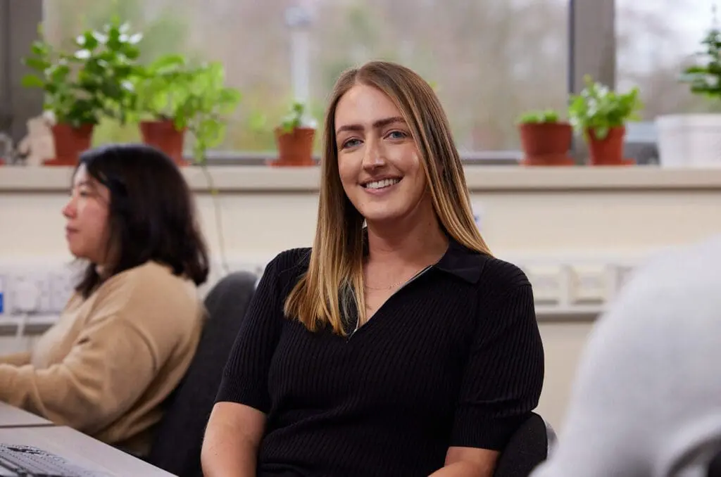 Woman smiling at desk in office with plants on windowsill; another person works at a computer nearby.