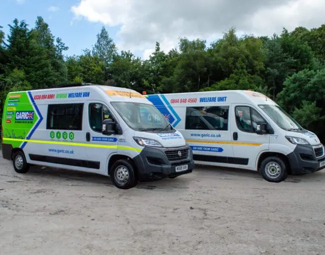 Two white welfare vans with colorful graphics are parked on a dirt lot, surrounded by green trees.