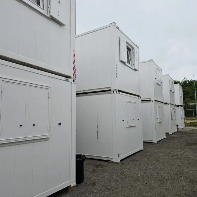 White portable cabins stacked in two rows on a gravel lot, with trees and fencing in the background.