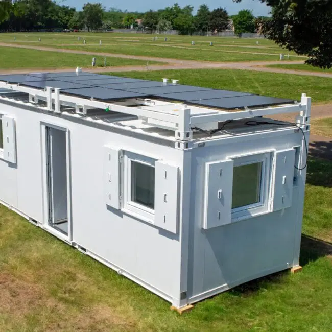 White container home with solar panels on the roof, placed on grass in an open, green field.