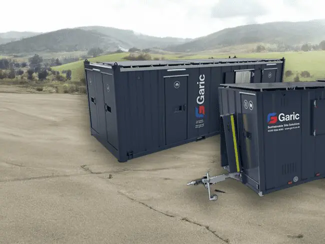 Two dark blue Garic portable cabins on a paved area with green hills and trees in the background.