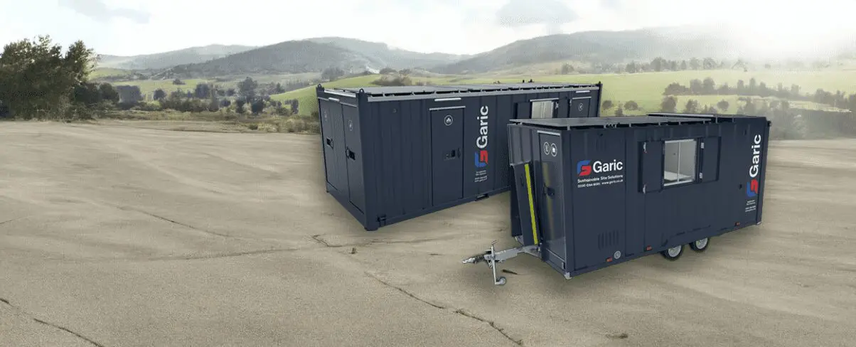 Two dark blue Garic portable cabins on a paved area with green hills and trees in the background.