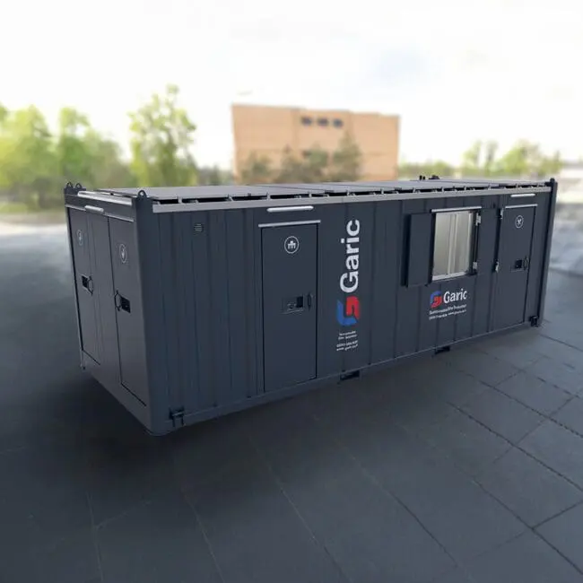 A dark grey Eco Cabin+ 24 portable site cabin by Garic sits on a paved outdoor area with blurred trees and a building in the background.