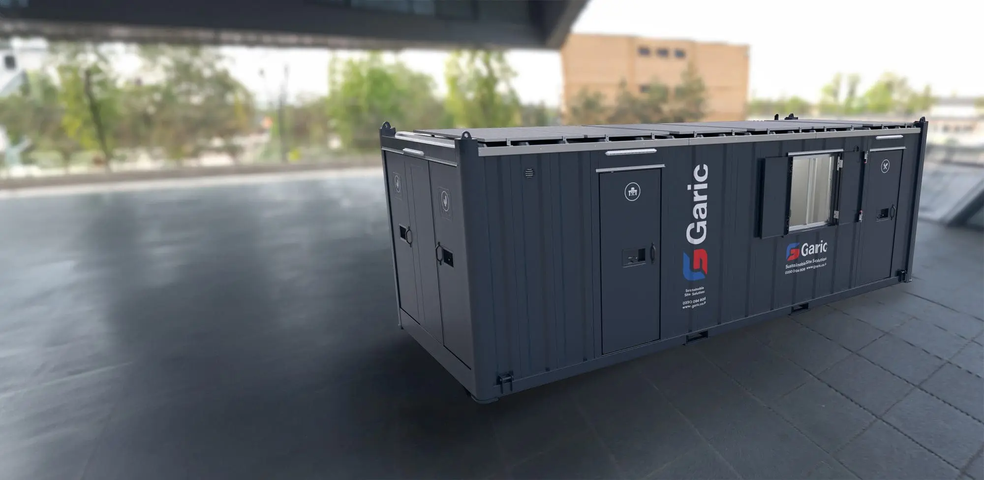 An Eco Cabin+ 24, large and dark gray, is positioned on an empty paved area with trees in the background.