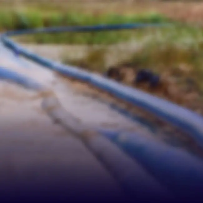 Blurry image of a pipe or hose on muddy ground, surrounded by grass and vegetation outdoors.