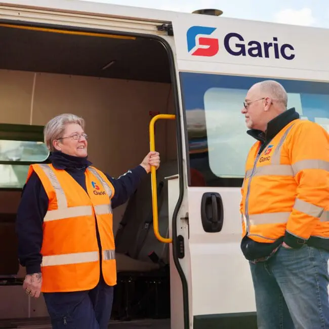 Two workers in orange safety vests stand by a Garic van, talking near the open side door.
