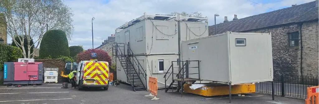Stacked portable cabins and a parked utility van in a lot, with people working nearby under a cloudy sky.