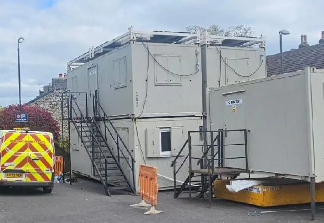 Stacked portable cabins and a parked utility van in a lot, with people working nearby under a cloudy sky.