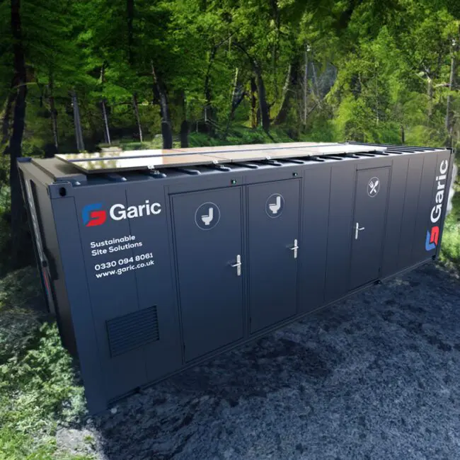A portable restroom unit by Garic is placed in a forest with sunlight filtering through the trees.