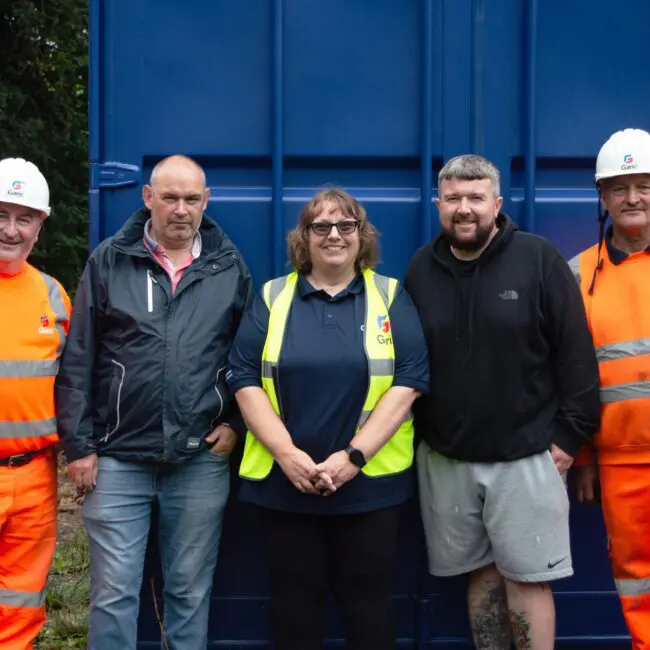 Five people, including two in orange workwear, pose and smile in front of a blue container outdoors.