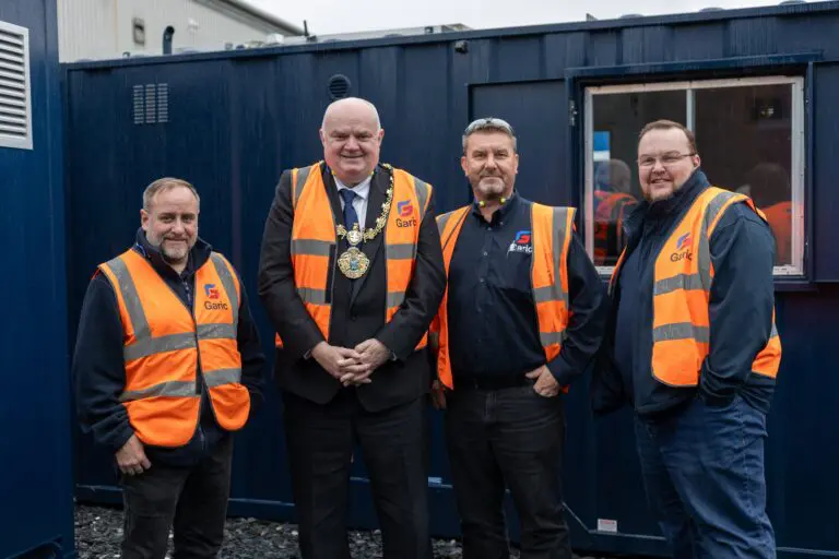 Four men in orange safety vests stand smiling in front of a blue portable building.