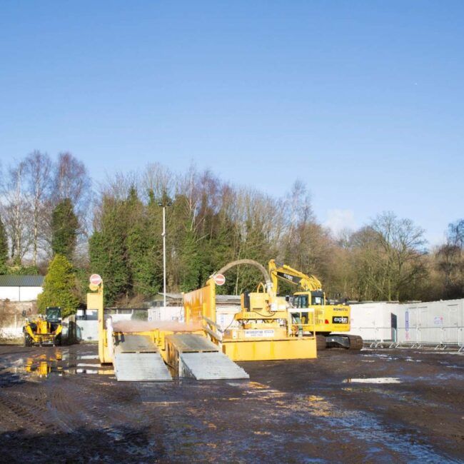 A construction site with workers, machinery, portable buildings, and muddy ground under a clear blue sky.