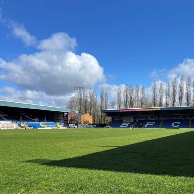 Empty football stadium with green grass, blue seats, and clear sky with fluffy clouds.