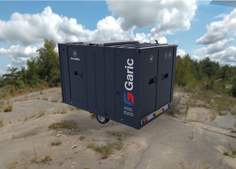 A dark gray Armadillo 12 Eco portable unit sits on cracked concrete, with grass and a cloudy sky in the background.