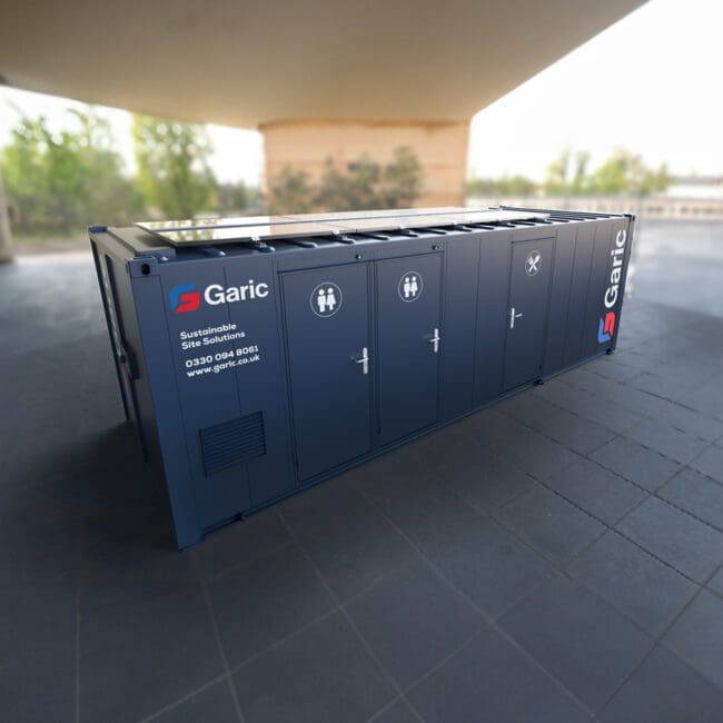 A blue Combi Cabin 24 Eco portable restroom unit sits outdoors on a tiled surface under a large structure.