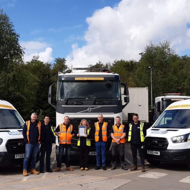 Eight people in safety vests stand in front of two vans and a truck in a parking lot on a sunny day.