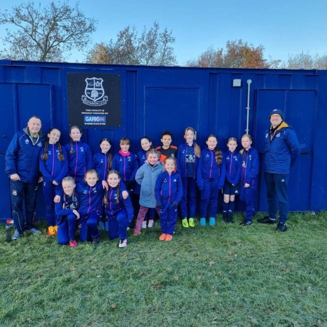 A girls soccer team and their coaches pose and smile in front of a blue container on a grassy field.